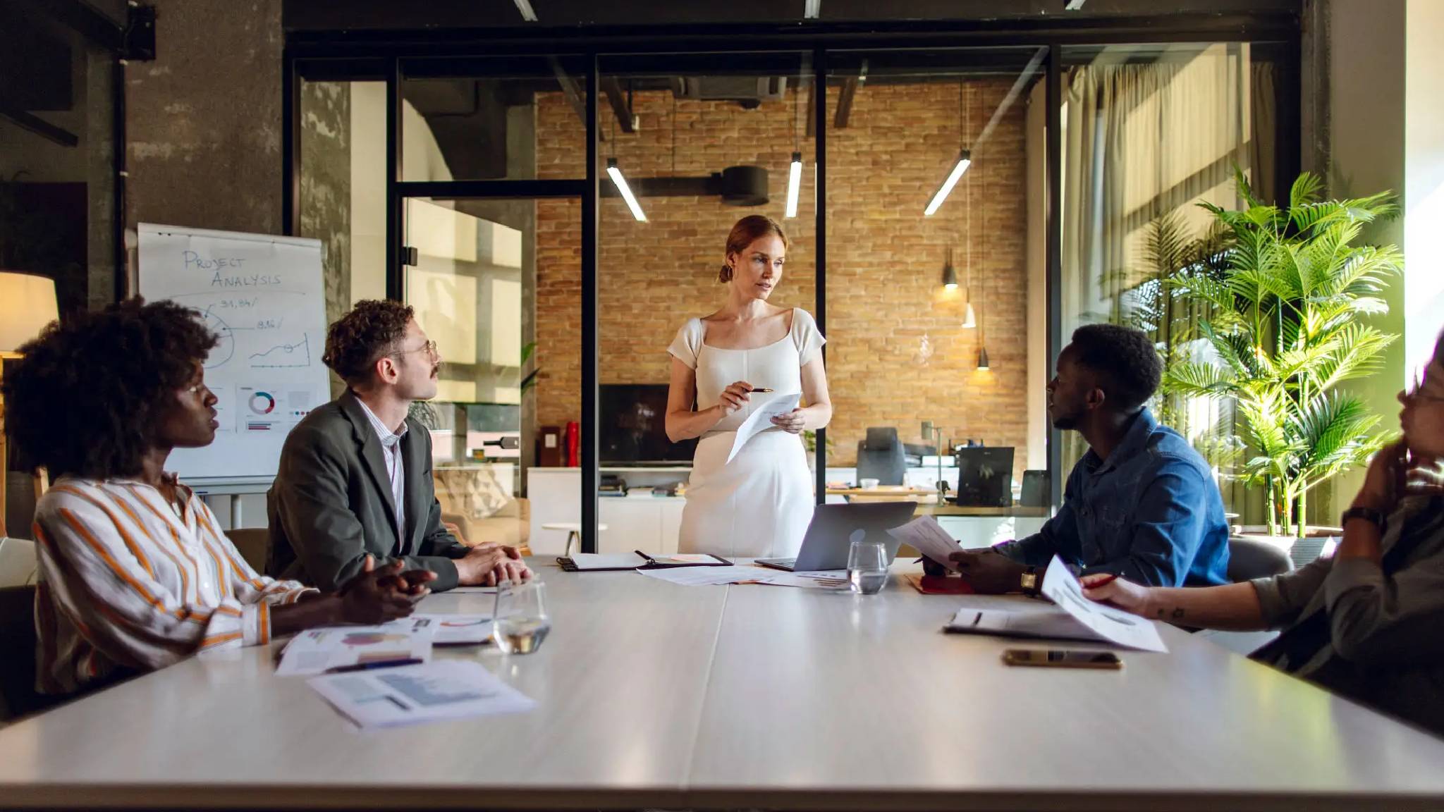 A woman stands confidently at a conference table, holding papers, addressing four attentive colleagues in a modern office with a brick wall backdrop